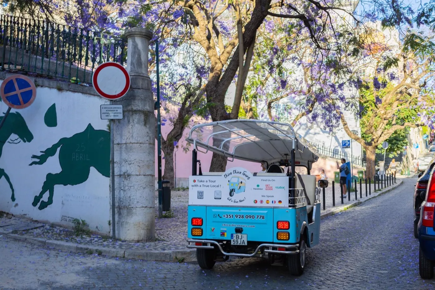 A tuk tuk under jacarandas next to a mural